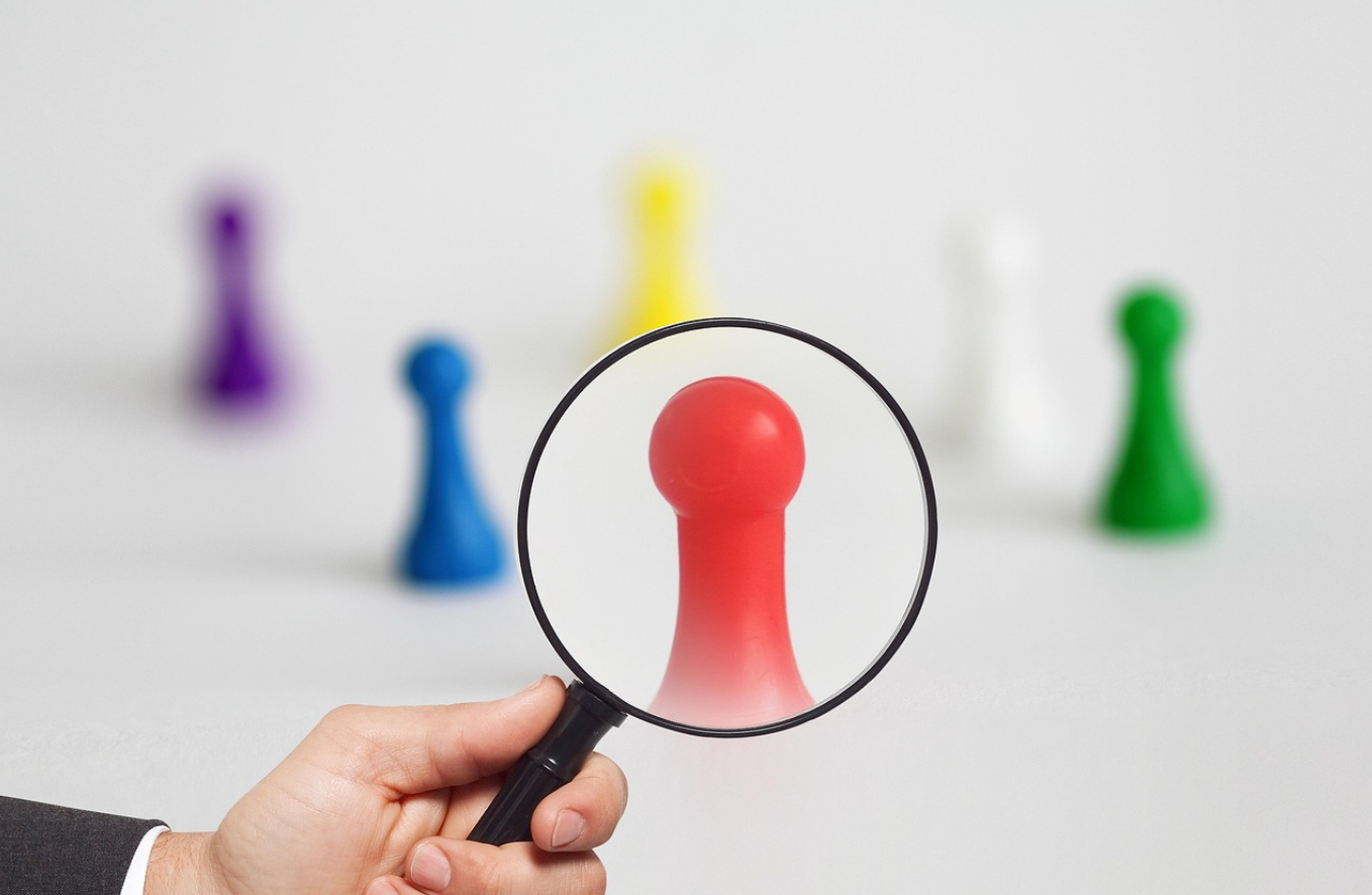 Multicoloured pieces from a game like Cluedo on a white background, with a person holding a magnifying glass up to the red piece.
